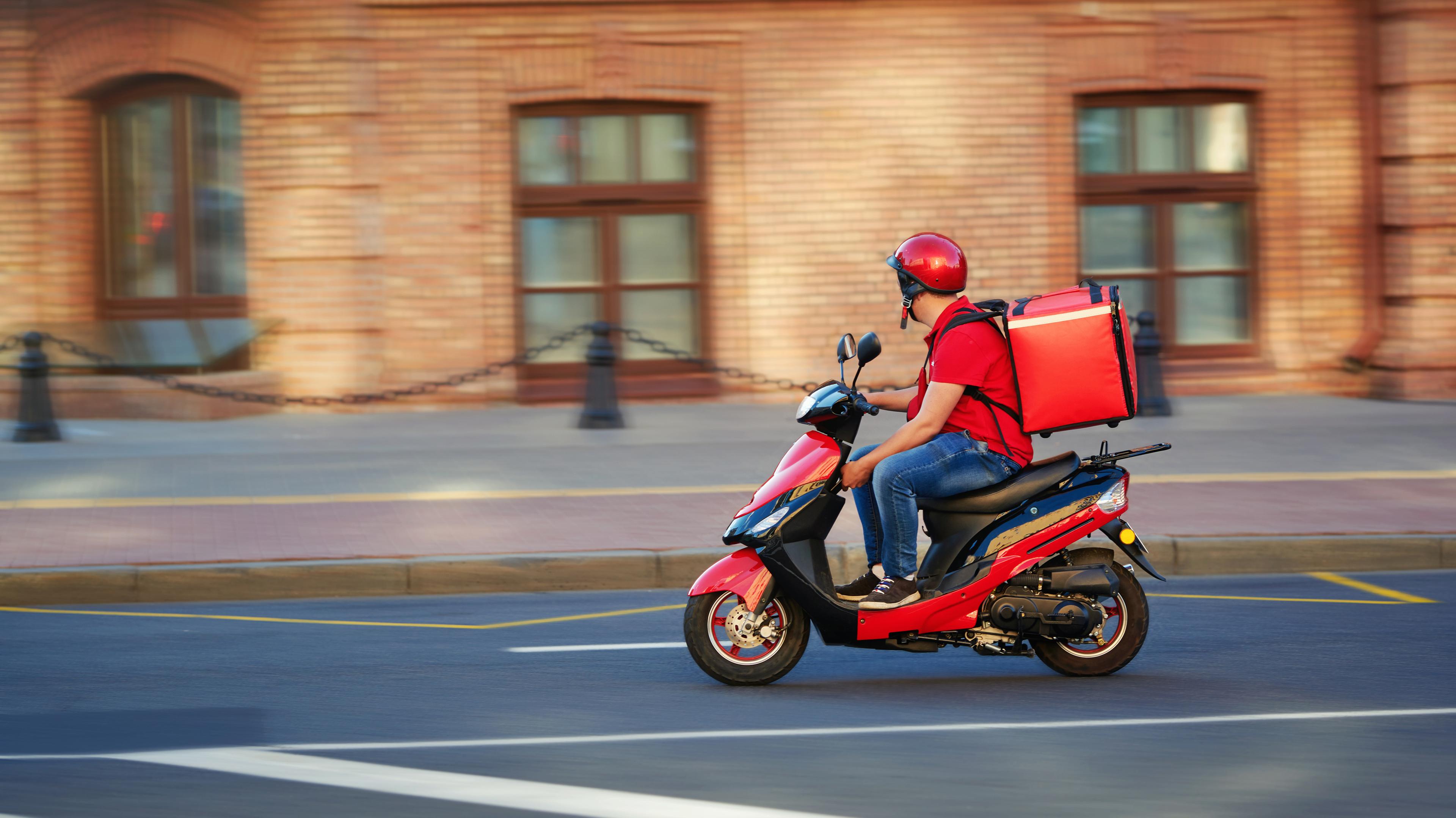 Ein Lieferdienstfahrer fährt mit seinem Moped und seiner roten Tragetasche auf dem Rücken durch die Straßen.