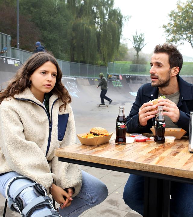 "Bettys Diagnose - Am Limit": Emily Hansen (Nina Kaiser) und Dr. Frank Stern (Max Alberti) sitzen an einem Tisch an einer Skateboard Bahn und essen Burger.
