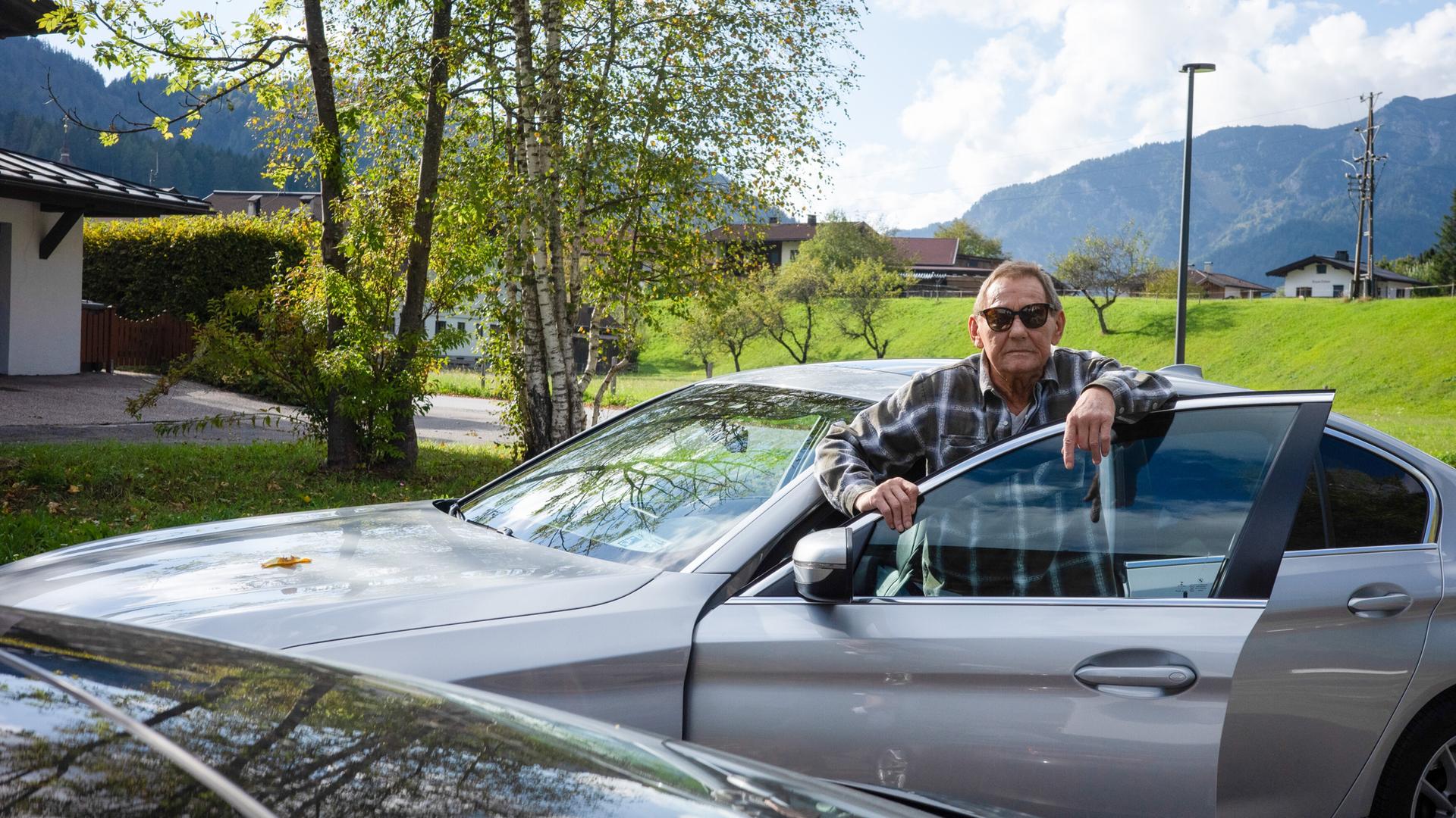 Das Bild zeigt einen älteren Mann, der an einem silbernen Auto steht. Er trägt eine kariierte Jacke und eine Sonnenbrille. Der Mann hat eine entspannte Haltung und lehnt an der offenen Tür des Fahrzeugs.   Im Hintergrund sind grüne Wiesen und Bäume zu sehen, die von der herbstlichen Färbung geprägt sind. Einige Häuser sind leicht unscharf im Hintergrund zu erkennen, und eine Berglandschaft ist ebenfalls sichtbar. Der Himmel ist blau mit einigen Wolken. Der gesamte Eindruck vermittelt eine ruhige ländliche Umgebung.