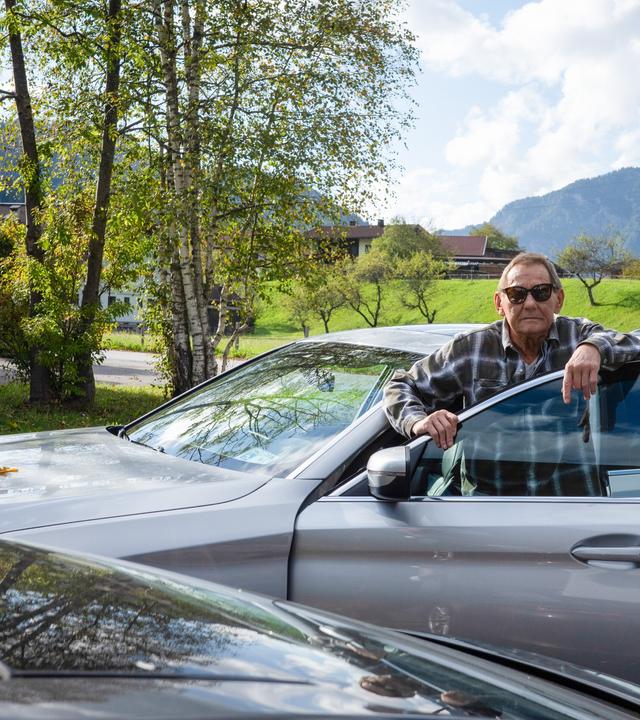 Das Bild zeigt einen älteren Mann, der an einem silbernen Auto steht. Er trägt eine kariierte Jacke und eine Sonnenbrille. Der Mann hat eine entspannte Haltung und lehnt an der offenen Tür des Fahrzeugs.   Im Hintergrund sind grüne Wiesen und Bäume zu sehen, die von der herbstlichen Färbung geprägt sind. Einige Häuser sind leicht unscharf im Hintergrund zu erkennen, und eine Berglandschaft ist ebenfalls sichtbar. Der Himmel ist blau mit einigen Wolken. Der gesamte Eindruck vermittelt eine ruhige ländliche Umgebung.