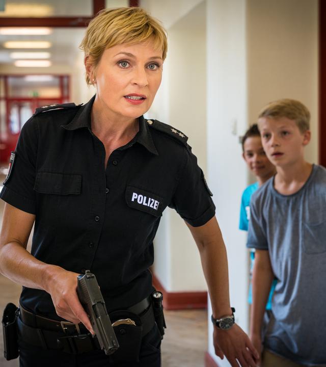 "Notruf Hafenkante - Amok": Melanie Hansen (Sanna Englund) geht mit schussbereiter Waffe über einen Schulflur. Seitlich hinter ihr stehen zwei Jungen in einer Klassenzimmertür. Melanie weist sie mit einem Handzeichen an, zurückzubleiben.