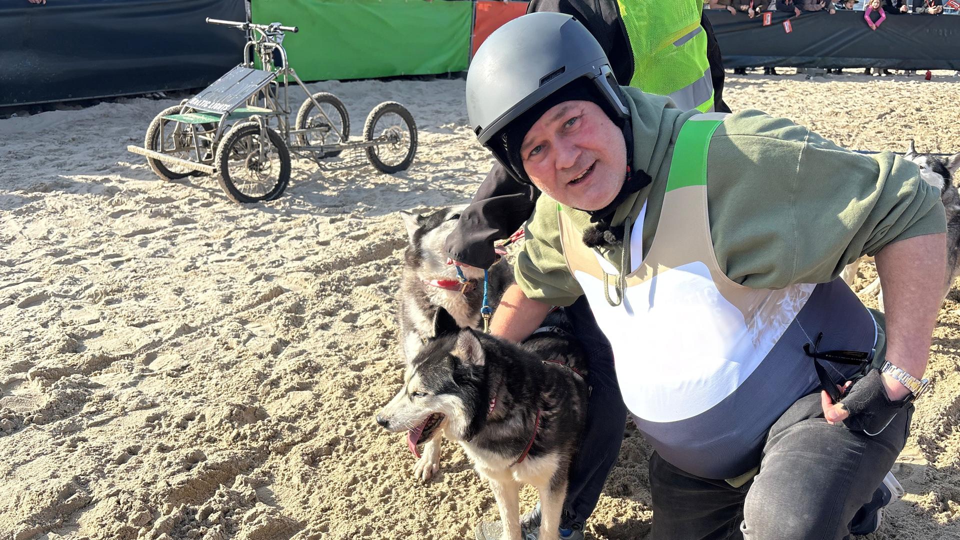 Waldi kniet neben zwei Schlittenhunden am Sandstrand und schaut in die Kamera.