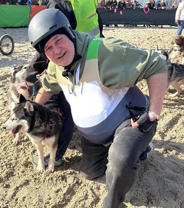 Waldi kniet neben zwei Schlittenhunden am Sandstrand und schaut in die Kamera.
