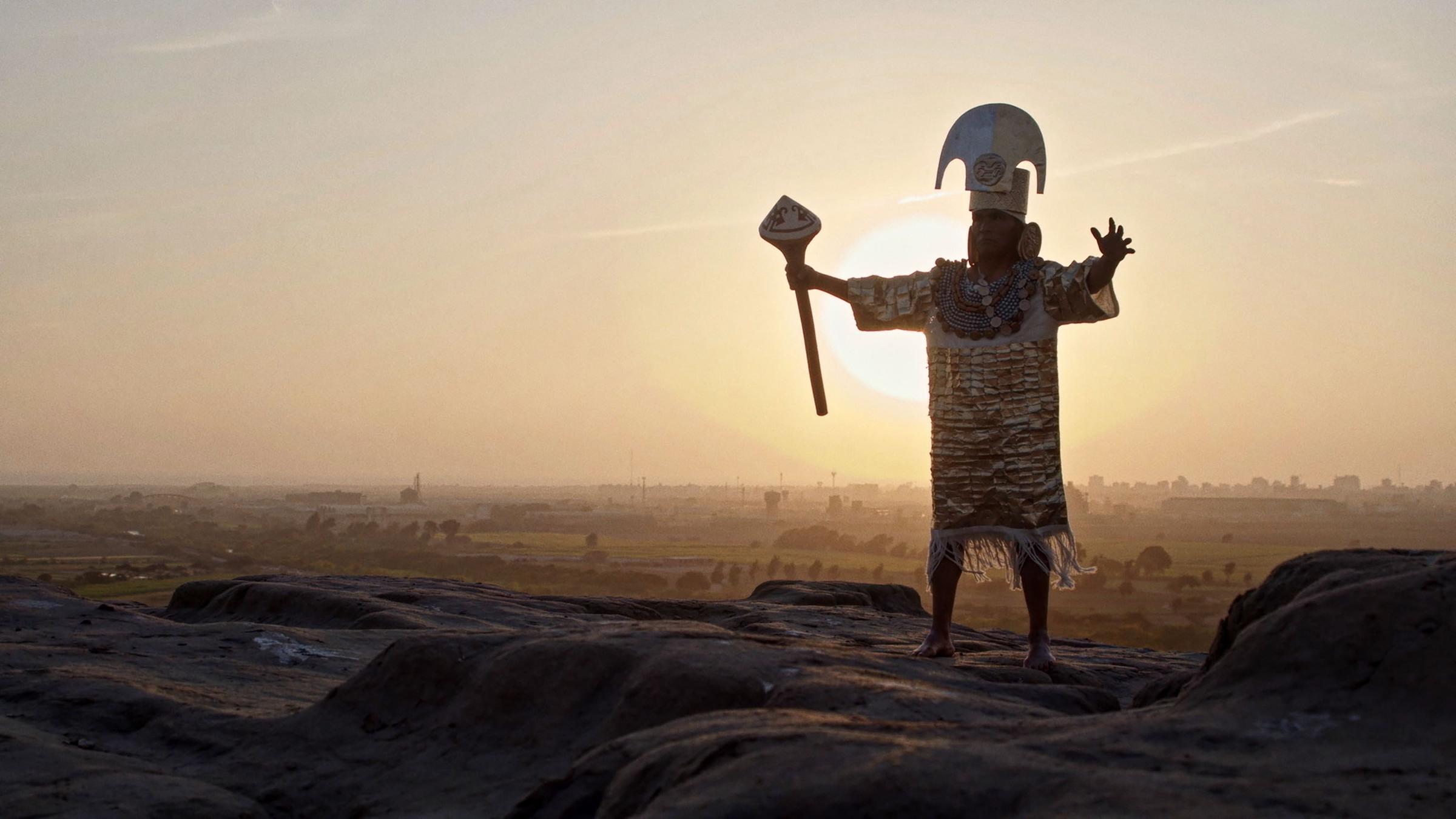 Ein Mann mit Kopfschmuck, festlichem Gewand und einer Art Zepter in der Hand steht mit ausgebreiteten Armen auf einem Hügel vor dem Sonnenuntergang.