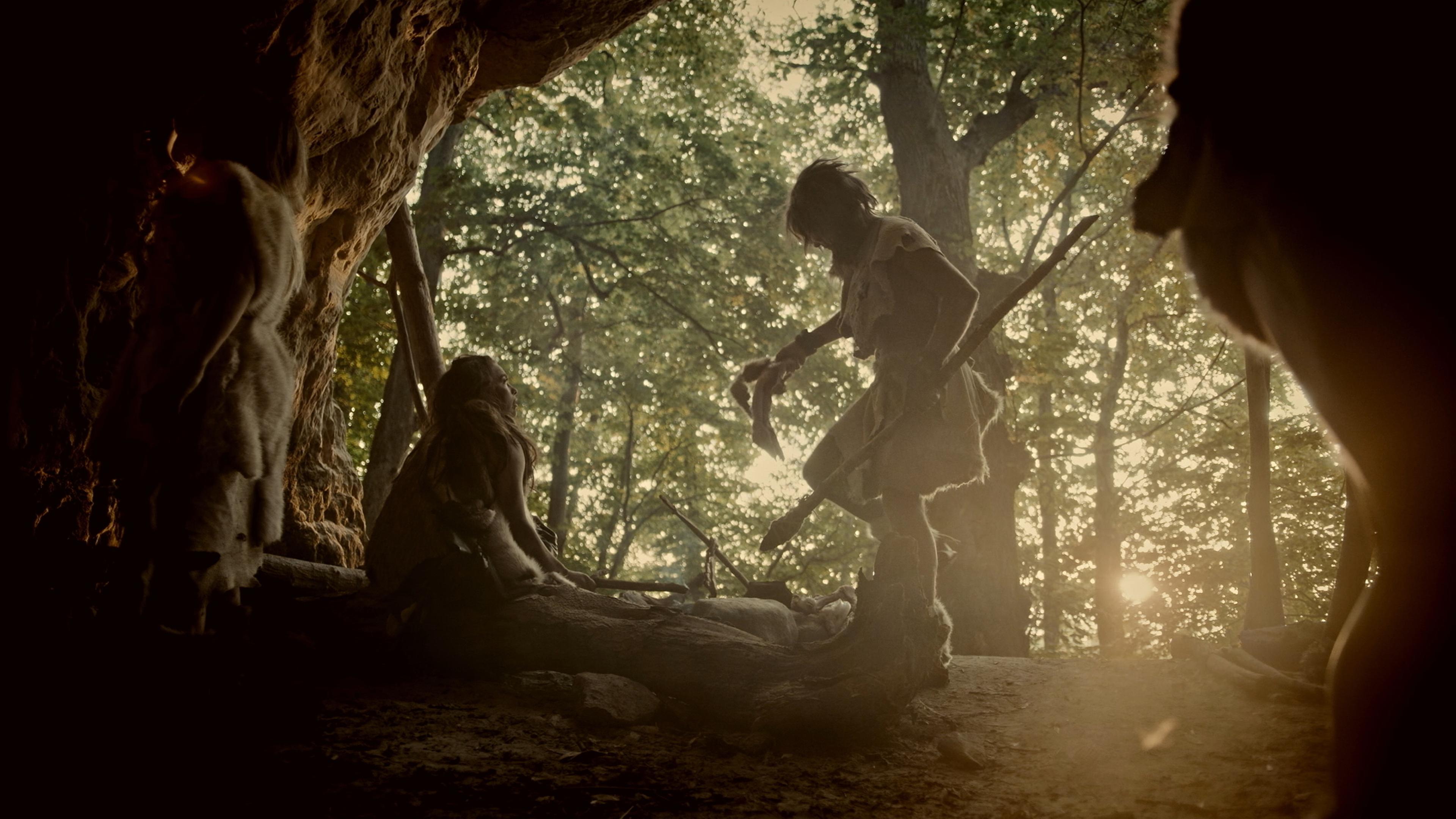 Blick aus dem Innern einer dunklen Höhle auf einen Wald. Im Höhleneingang sitzt eine Person neben einer Feuerstelle. Daneben steht ein in Fell gekleideter Mensch mit steinzeitlichem Speer.