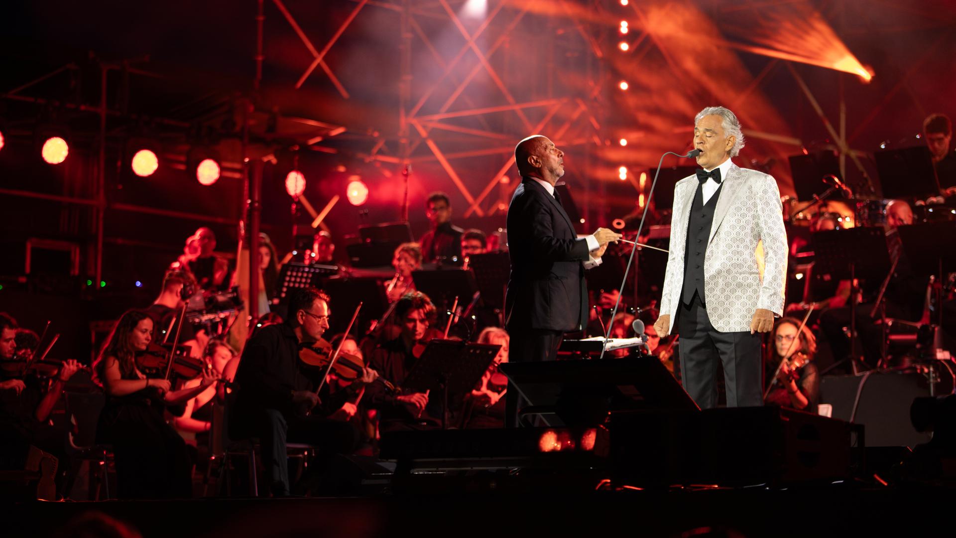 Andrea Bocelli (rechts) steht auf einer Bühne und singt in ein Mikrofon. Links von ihm steht der Dirigent und im Hintergrund ist ein Orchester zu sehen.