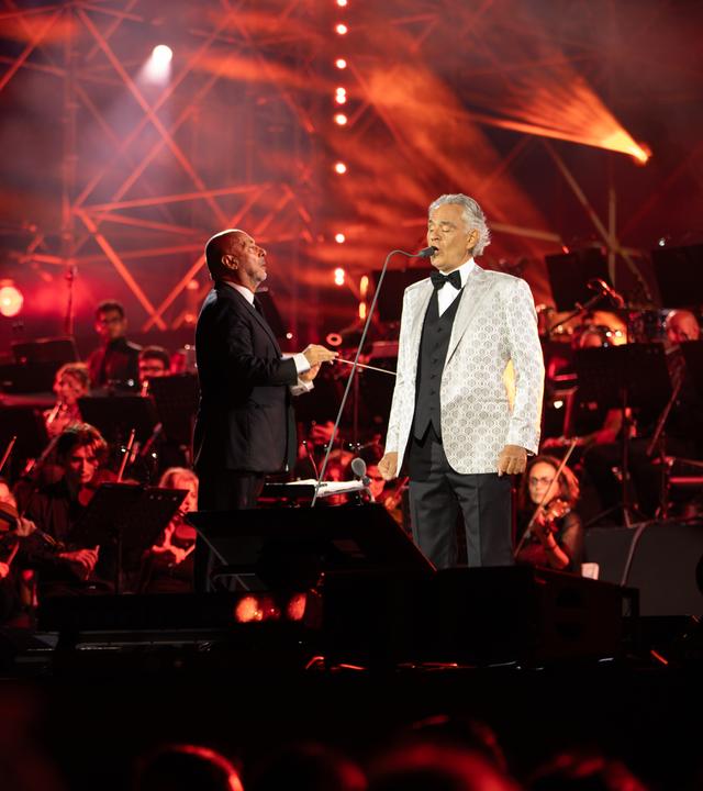 Andrea Bocelli (rechts) steht auf einer Bühne und singt in ein Mikrofon. Links von ihm steht der Dirigent und im Hintergrund ist ein Orchester zu sehen.