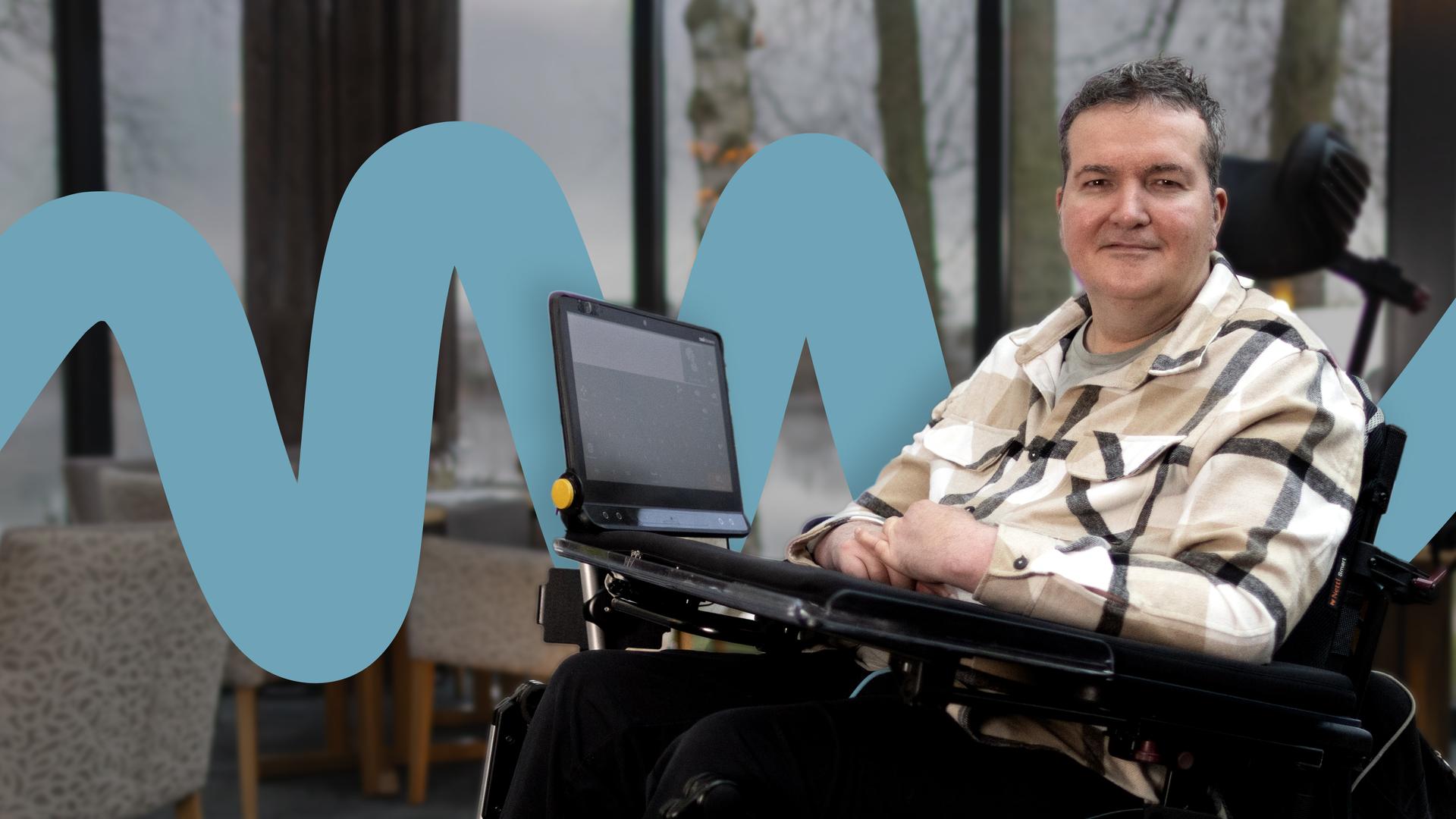 Andreas Pennings sitzt in einem elektrischen Rollstuhl an einem gedeckten Restauranttisch vor großen Fenstern mit Blick auf einen Fluss und winterliche Bäume. Auf seinem Rollstuhltisch ist ein Monitor mit Augensteuerung befestigt. Er trägt ein hell kariertes Hemd und blickt ruhig in die Kamera. Im Hintergrund stehen weitere Tische mit Gläsern und Kerzenlicht.
