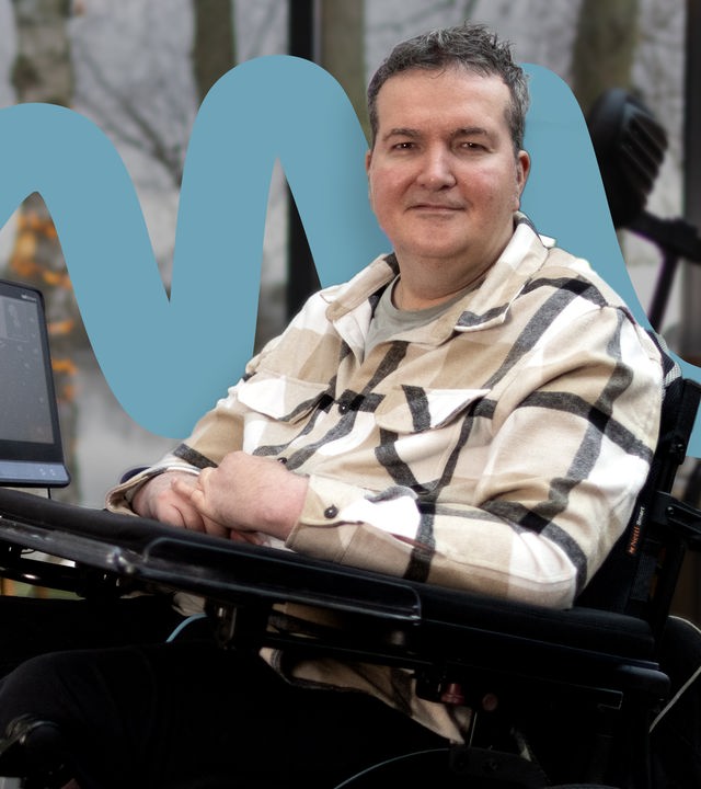 Andreas Pennings sitzt in einem elektrischen Rollstuhl an einem gedeckten Restauranttisch vor großen Fenstern mit Blick auf einen Fluss und winterliche Bäume. Auf seinem Rollstuhltisch ist ein Monitor mit Augensteuerung befestigt. Er trägt ein hell kariertes Hemd und blickt ruhig in die Kamera. Im Hintergrund stehen weitere Tische mit Gläsern und Kerzenlicht.