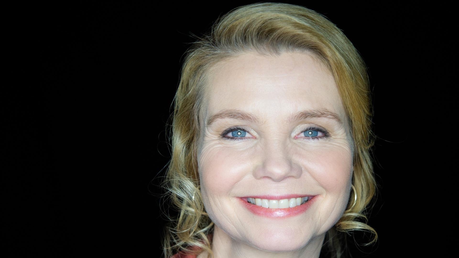 Porträt von Annette Frier vor schwarzem Hintergrund.Sie trägt eine rote Bluse und blickt strahlend in den Kamara.