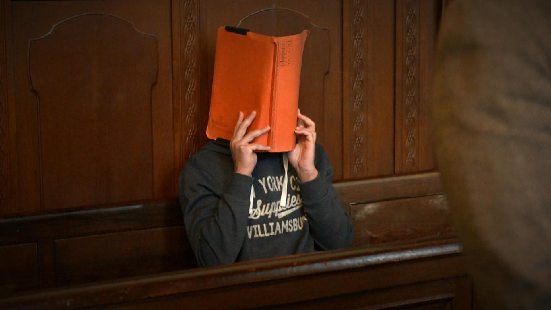 "Angeklagt! Mord oder Totschlag?": Ein Mann sitzt auf einer Bank in einem Gerichtssaal, vor sein Gesicht hält er eine orangefarbene Mappe.