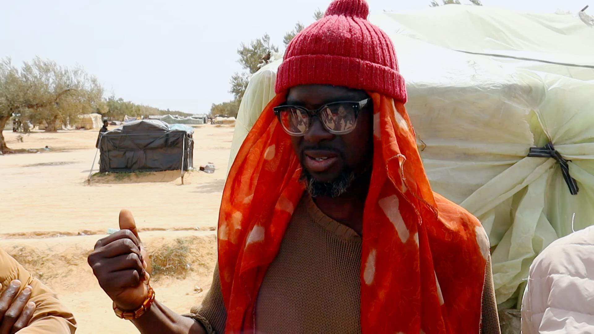 Der aus Nigeria geflüchtete Angelou Happyvidar steht in einem Camp für Geflüchtete nach der tunesischen Haftenstadt Sfax. Er trägt eine Sonnebrille, ein kurzärmliges Oberteil und ein dünnes Tuch unter einer Wollmütze.