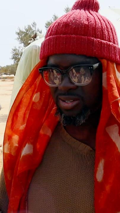 Der aus Nigeria geflüchtete Angelou Happyvidar steht in einem Camp für Geflüchtete nach der tunesischen Haftenstadt Sfax. Er trägt eine Sonnebrille, ein kurzärmliges Oberteil und ein dünnes Tuch unter einer Wollmütze.