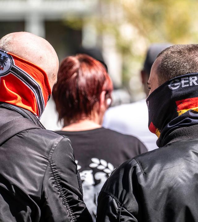 Foto zweier Männer in schwarzen Lederjacken von hinten. Einer ist kahlgeschoren und hat einen Schlauchschal mit Reichsflagge bis über die Nase hochgezogen. Der zweite ist kurzgeschoren und hat einen Schlauchschal mit Deutschlandfarben und dem Aufdruck Germany halb übers Gesicht gezogen.