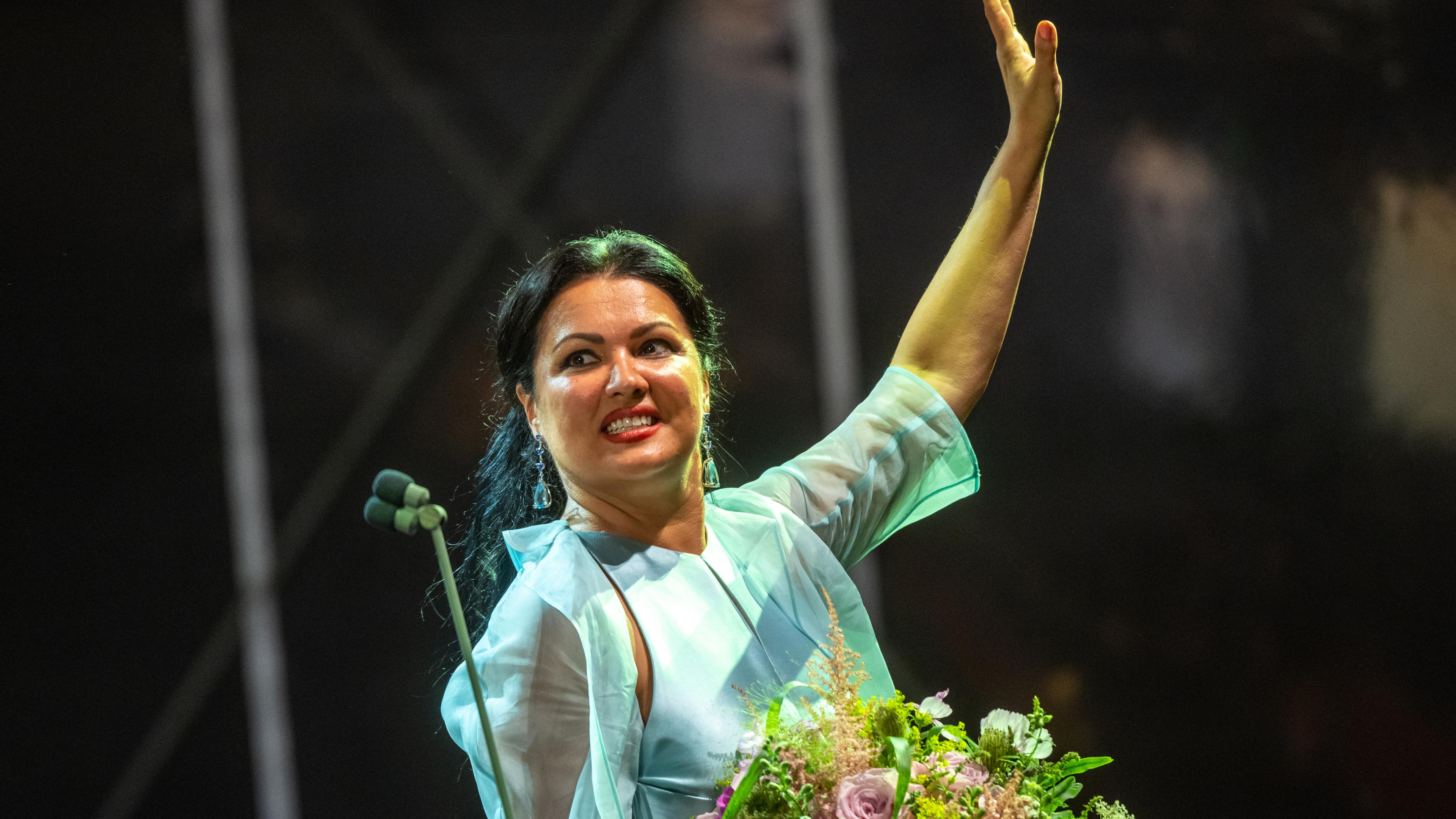 Die Sängerin Anna Netrebko hält, festlich gekleidet, einen Blumenstrauß. Die andere Hand hat sie zum Gruß gehoben.