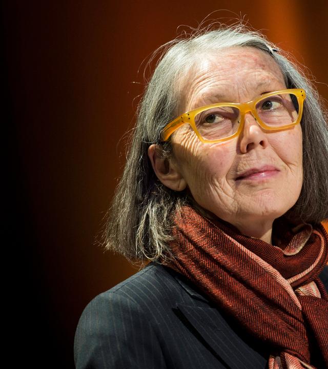 Porträtfoto einer älteren Frau mit schulterlangen grauen Haaren und einer Brille mit gelbem Kunststoffrahmen.