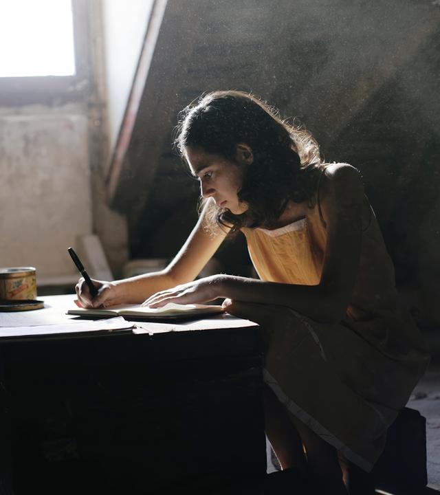 Das Bild in Schwarz-Weiß zeigt ein Mädchen an einem Schreibtisch, vor ihm liegt ein geöffnetes Buch und es hält einen Stift in der Hand. Das Mädchen trägt einen dunklen Pullover mit einer hellen Bluse darunter. Sie blickt neutral in die Kamera. Hinter ihr steht eine Topfpflanze.