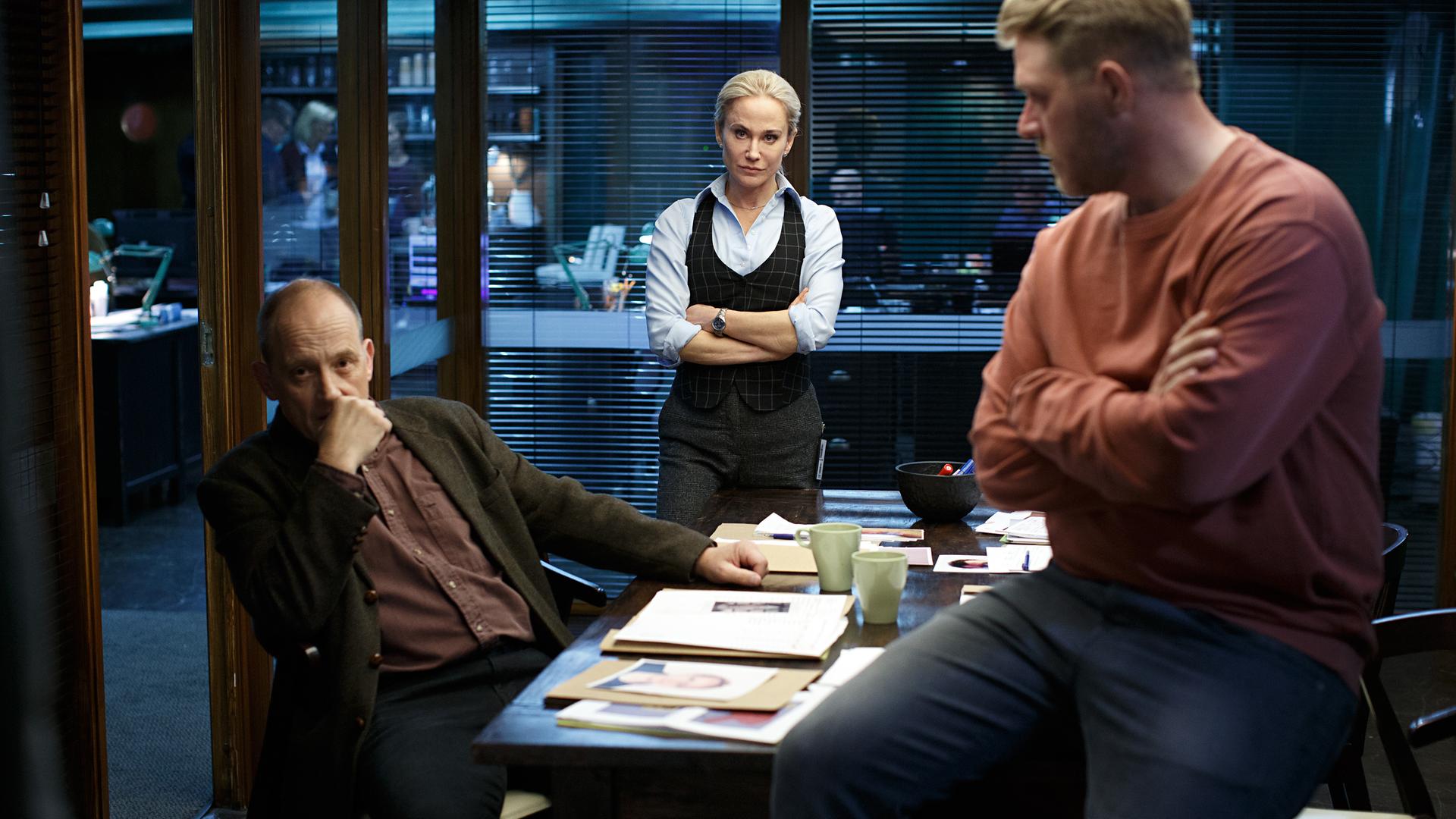 "Anne Holt: Der Mörder in uns": Der Ermittler Torbjörn Skoglund (Samuel Fröler) sitzt in einem Büro mit gläsernen Wänden an seinem Schreibtisch. Seine Kollegen Jessica Östlund (Anja Lundqvist) und Ingvar Nyman (Henrik Norlén) stehen mit verschränkten Armen vor ihm. Alle drei sehen nachdenklich aus.