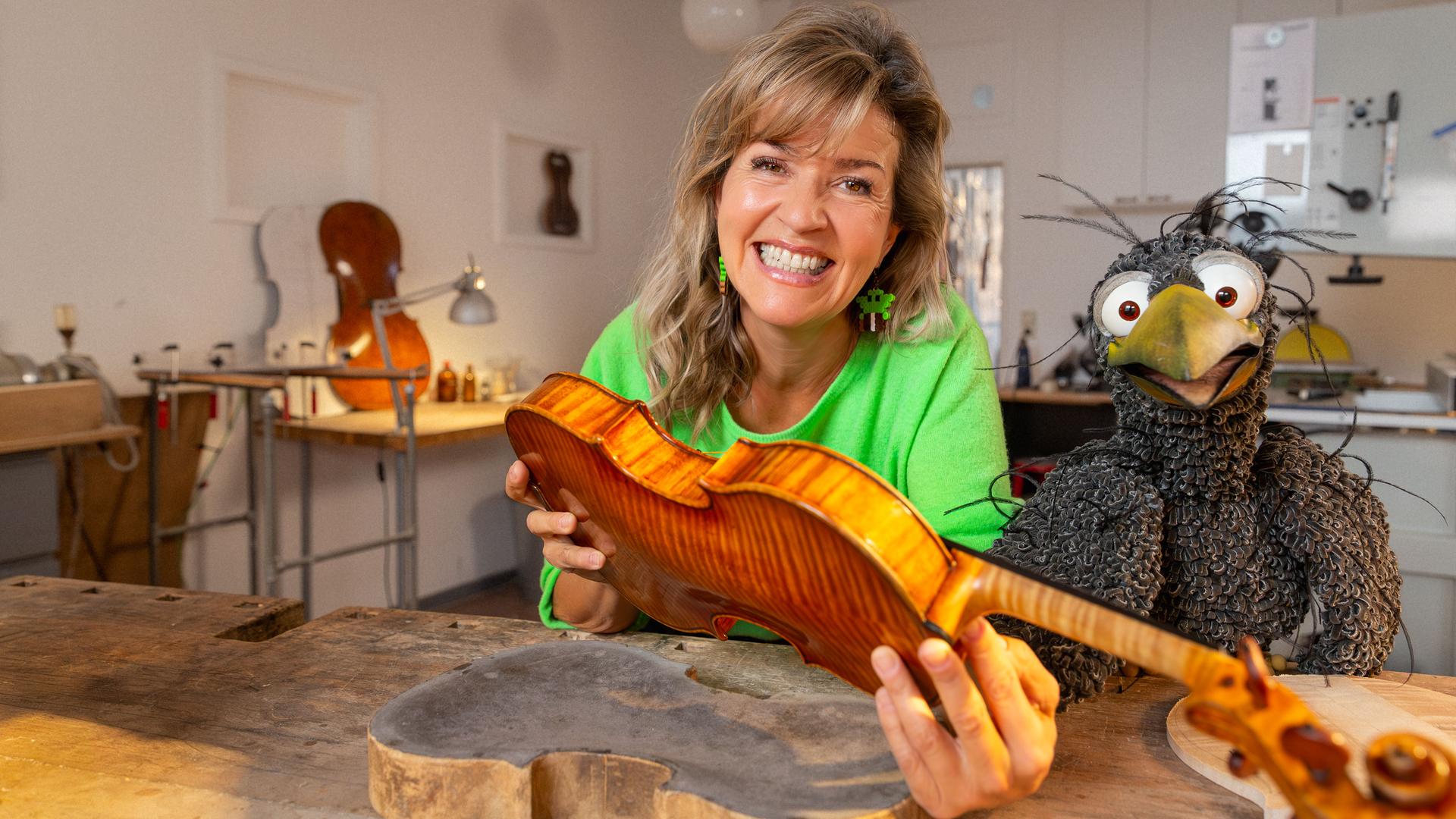 Anne-Sophie Mutter hält eine unfertige Geige in den Händen, neben ihr am Geigenbauer-Tisch hockt Rudi. Beide schauen freudig in die Kamera.