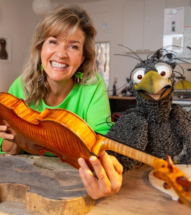Anne-Sophie Mutter hält eine unfertige Geige in den Händen, neben ihr am Geigenbauer-Tisch hockt Rudi. Beide schauen freudig in die Kamera.