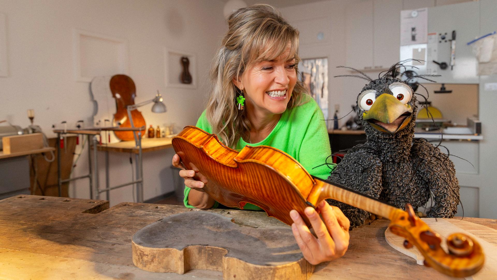 Anne-Sophie Mutter hält eine unfertige Geige in den Händen, neben ihr am Geigenbauer-Tisch hockt Rudi. Beide schauen freudig.