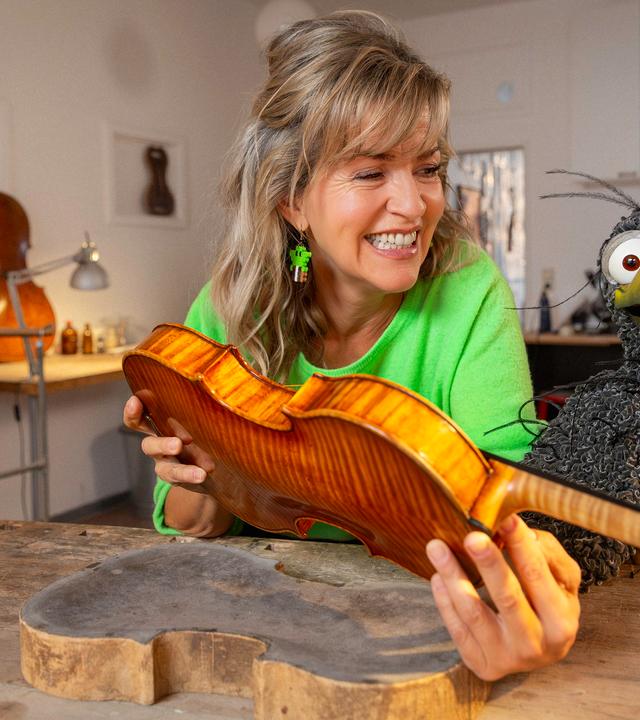 Anne-Sophie Mutter hält eine unfertige Geige in den Händen, neben ihr am Geigenbauer-Tisch hockt Rudi. Beide schauen freudig.