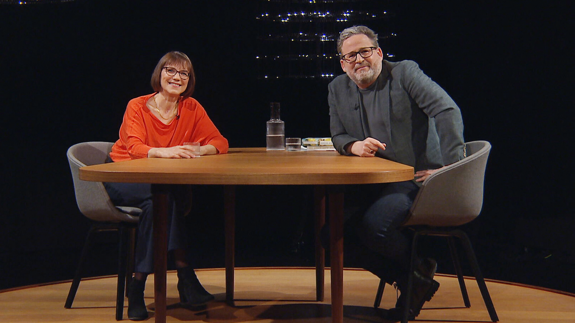 Zwei Personen sitzen sich in einem dunklen Studio an einem Holztisch gegenüber; links Annette Kehnel, rechts Moderator Wolfram Eilenberger.