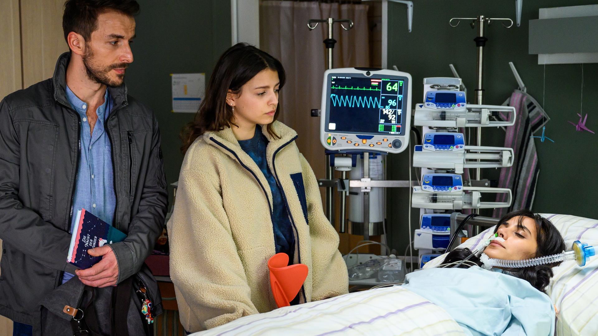 Dr. Frank Stern (Max Alberti) steht mit Emily Hansen (Nina Kaiser) am Krankenbett von ihrer Mutter Paula Hansen (Maya Henselek), die im Koma liegt. Emily und Dr. Stern schauen besorgt auf die Patientin.