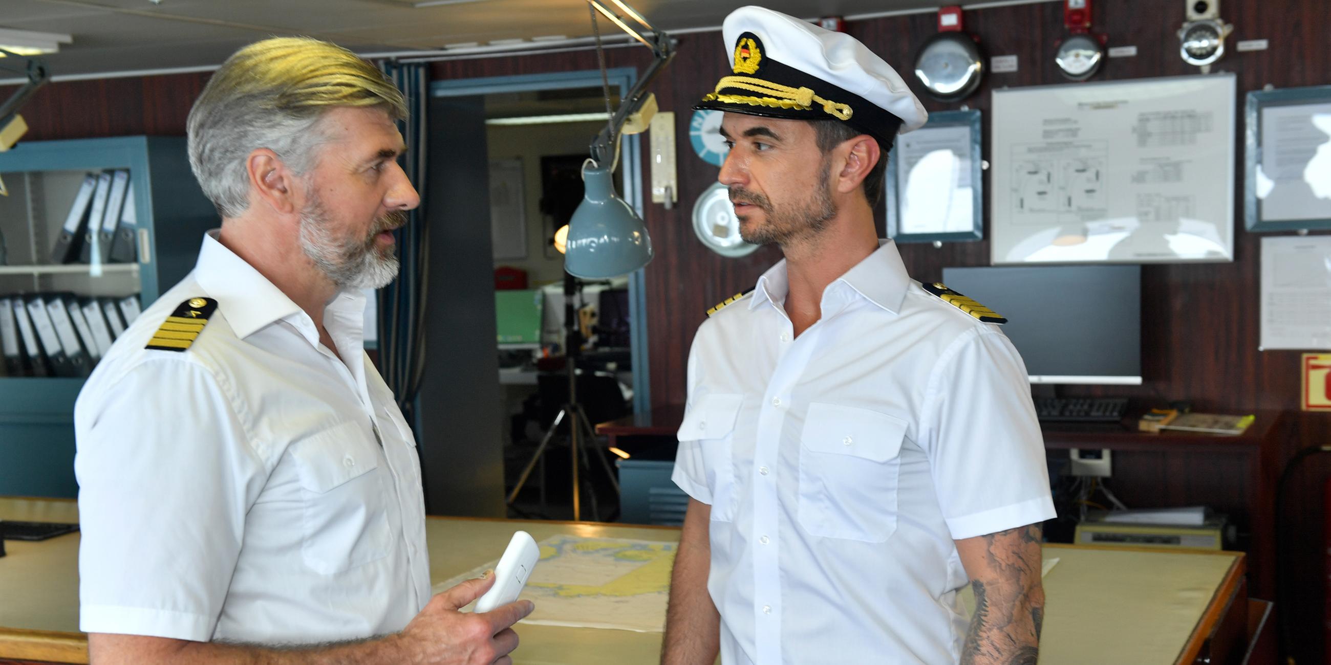 Staff-Kapitän Martin Grimm (Daniel Morgenroth) und Kapitän Max Parger (Florian Silbereisen) stehen auf der Brücke des Schiffs und schauen sich gegenseitig an.