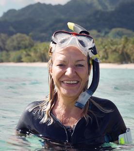 Die Systemforscherin Antje Boetius steht lächelnd im Wasser. Sie trägt eine Taucherbrille auf dem Kopf. Hinter ihr liegt eine bewaldete Hügelkette mit einem Sandstrand.