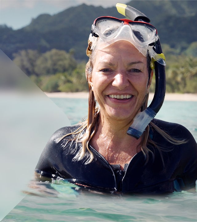 Die Systemforscherin Antje Boetius steht lächelnd im Wasser. Sie trägt eine Taucherbrille auf dem Kopf. Hinter ihr liegt eine bewaldete Hügelkette mit einem Sandstrand.