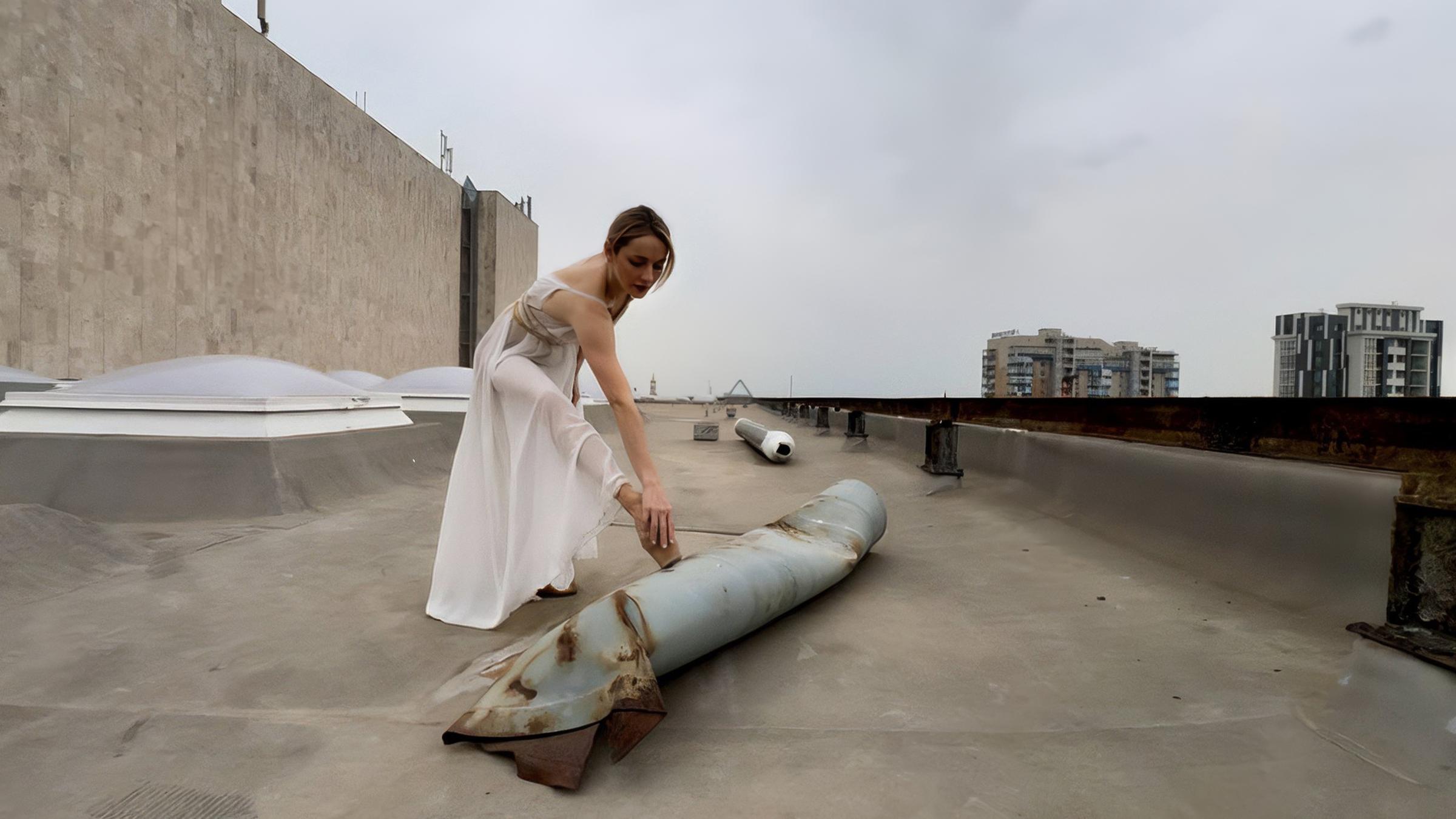 Eine junge Ballerina in durchsichtigem langem Gewand steht auf dem Dach eines Hochhausses und dehnt sich, indem sie einen Fuß auf den Überresten einer Rakete aufstellt.