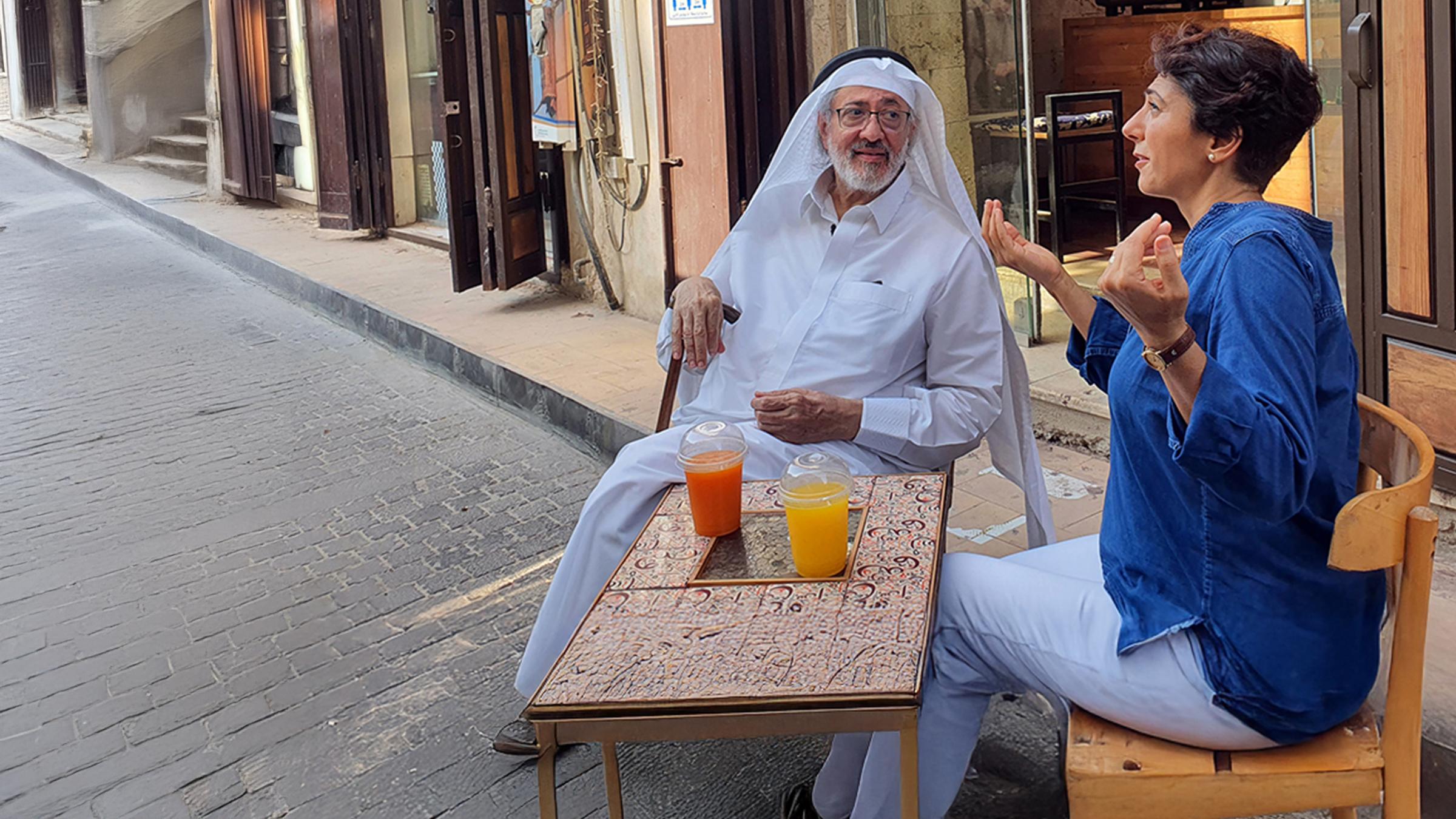 Golineh Atai mit dem Künstler Hisham Binjabi in Al Balad, der Altstadt von Dschidda in Saudi-Arabien. Sie sitzen an einem kleinen Tisch, auf dem Getränke vor ihnen stehen. Golineh Atai hat gestikulierend die Hände erhoben und schaut leicht nach oben. Hisham Binjabi lehnt auf seinem Stock und schaut Golineh Atai aufmerksam an.