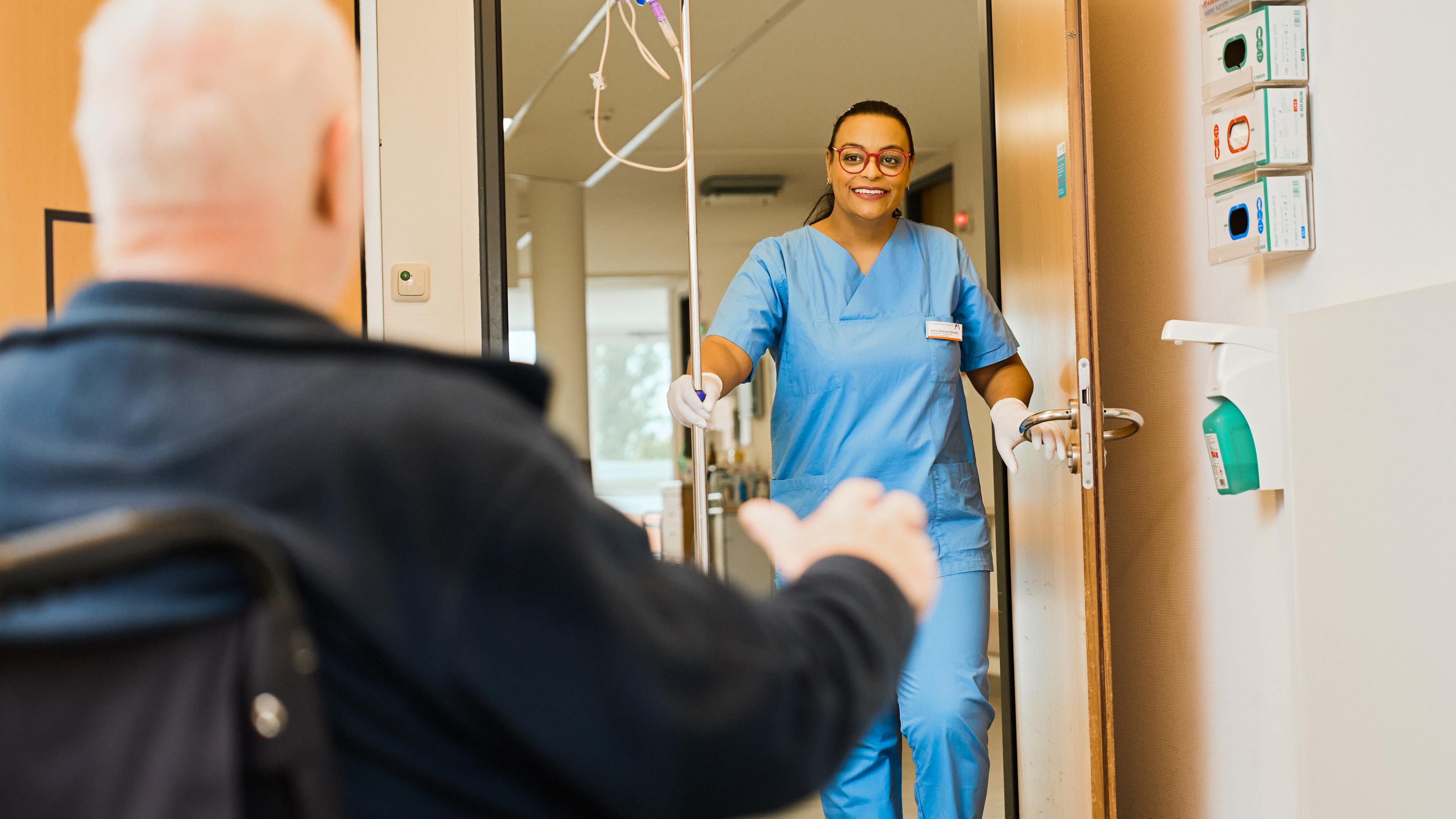 Im Krankenhaus: Elaine, die Pflegekraft steht lächelnd in der offenen Tür. Ein älterer Mann im sitzt, mit dem Rücken zur Kamera, im Rollstuhl in einem Zimmer.