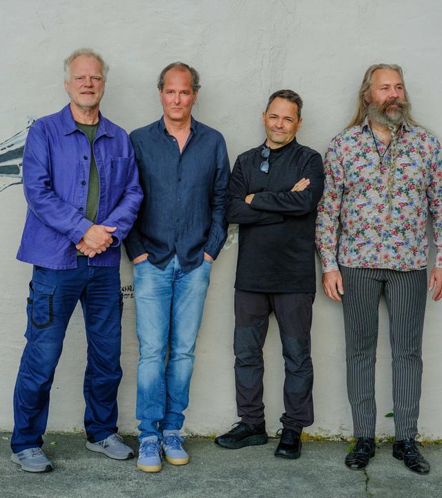 Vier Musiker stehen nebeneinander vor hellem Hintergrund; die Gruppe ist vollständig zu sehen und locker angeordnet.