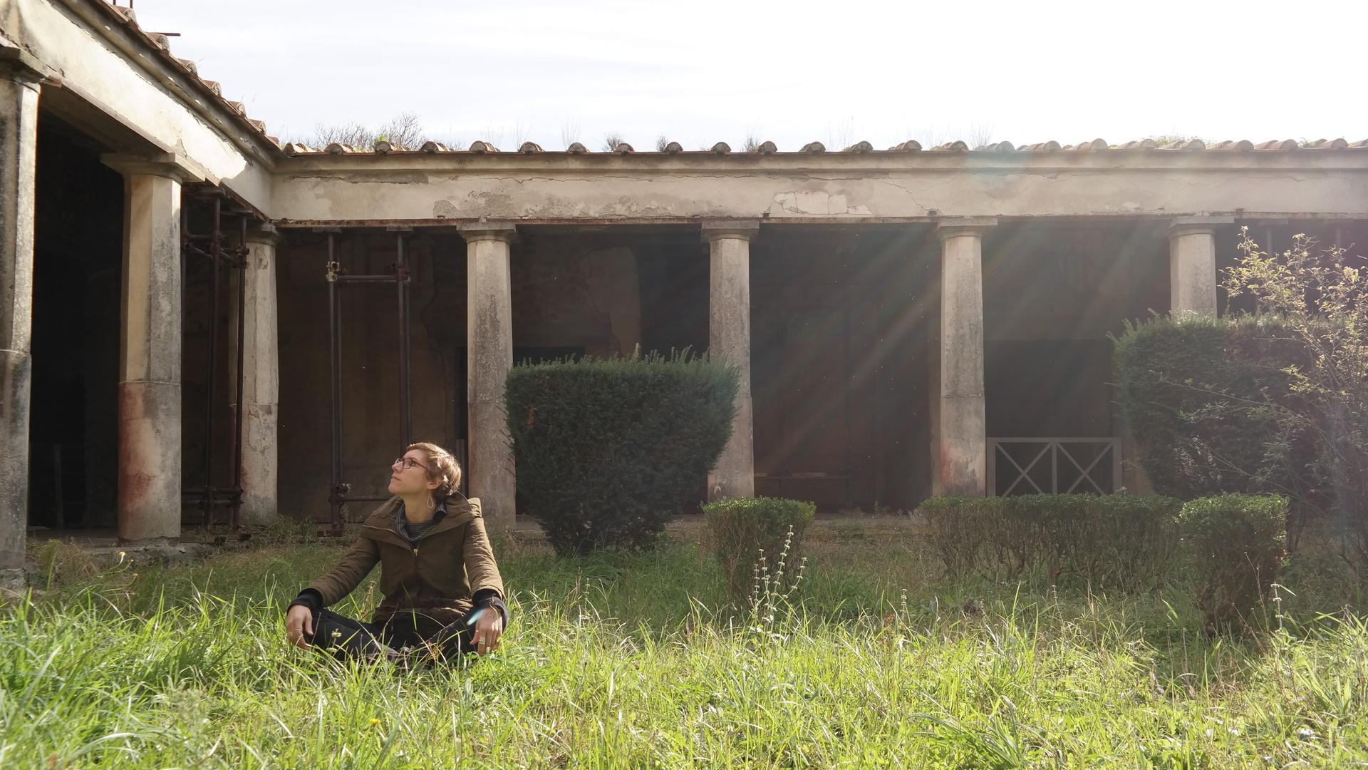 Eine junge Frau sitzt auf einer Wiese vor einem antiken Gebäude