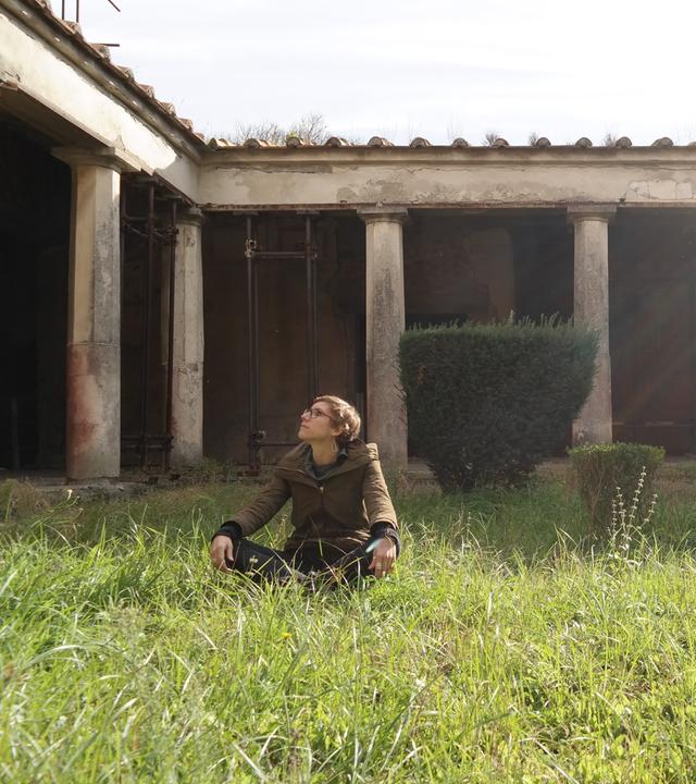 Eine junge Frau sitzt auf einer Wiese vor einem antiken Gebäude