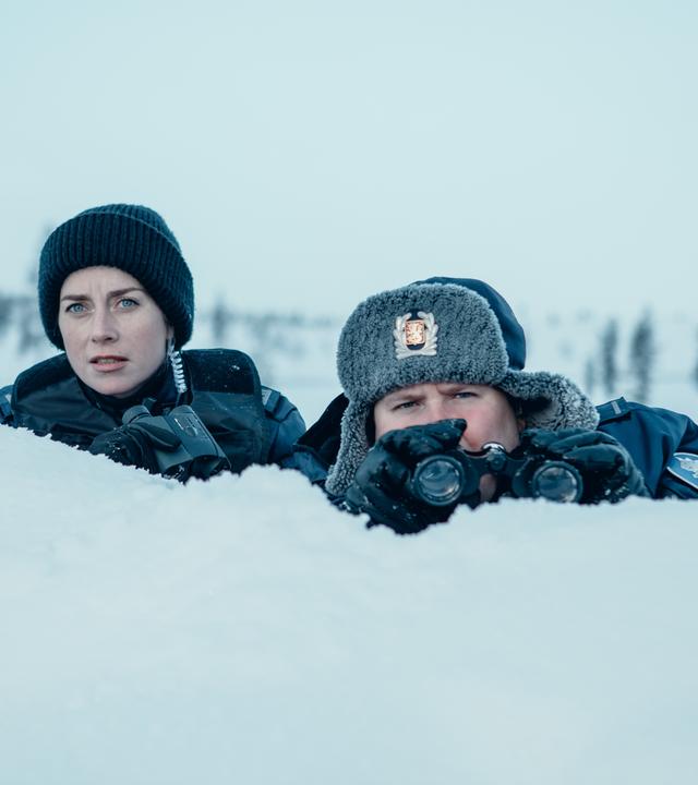"Arctic Circle - Der unsichtbare Tod (6)": Nina Kautsalo (Iina Kuustonen) und ihr Kollege Niilo Aikio (Janne Kataja) sitzen halb hinter einem schneebedeckten Hügel und schauen konzentriert nach vorne. Beide haben Ferngläser in der Hand.