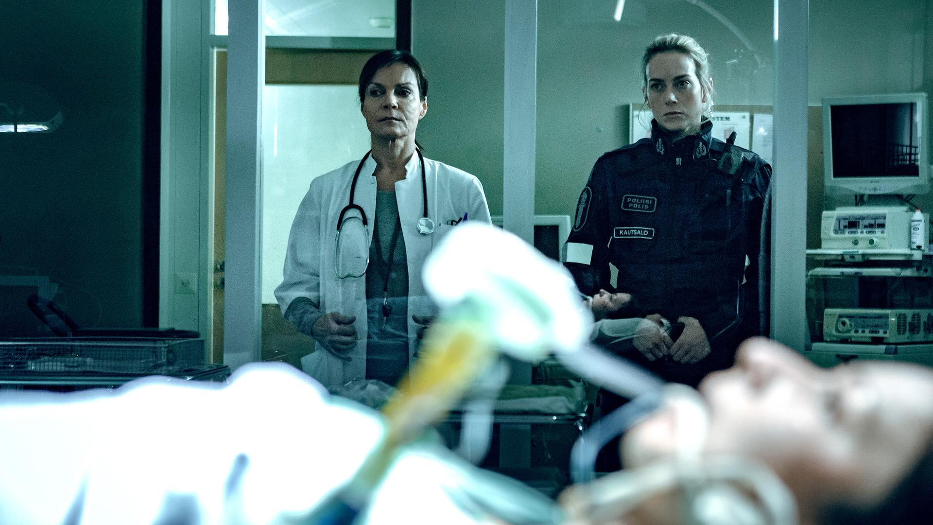 Die Ärztin Satu Kela (Lena Meriläinen) und die Polizistin Nina Kautsalo (Iina Kuustonen) stehen nebeneinander hinter einer Glasscheibe und schauen auf die schwer verletzte Irina (Anna Ackerman), die in einem Krankenhausbett liegt.
