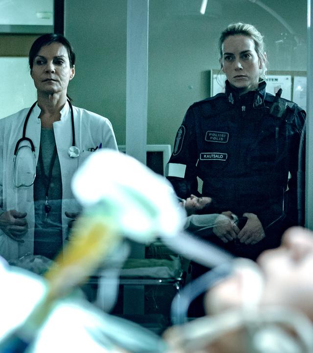 Die Ärztin Satu Kela (Lena Meriläinen) und die Polizistin Nina Kautsalo (Iina Kuustonen) stehen nebeneinander hinter einer Glasscheibe und schauen auf die schwer verletzte Irina (Anna Ackerman), die in einem Krankenhausbett liegt.