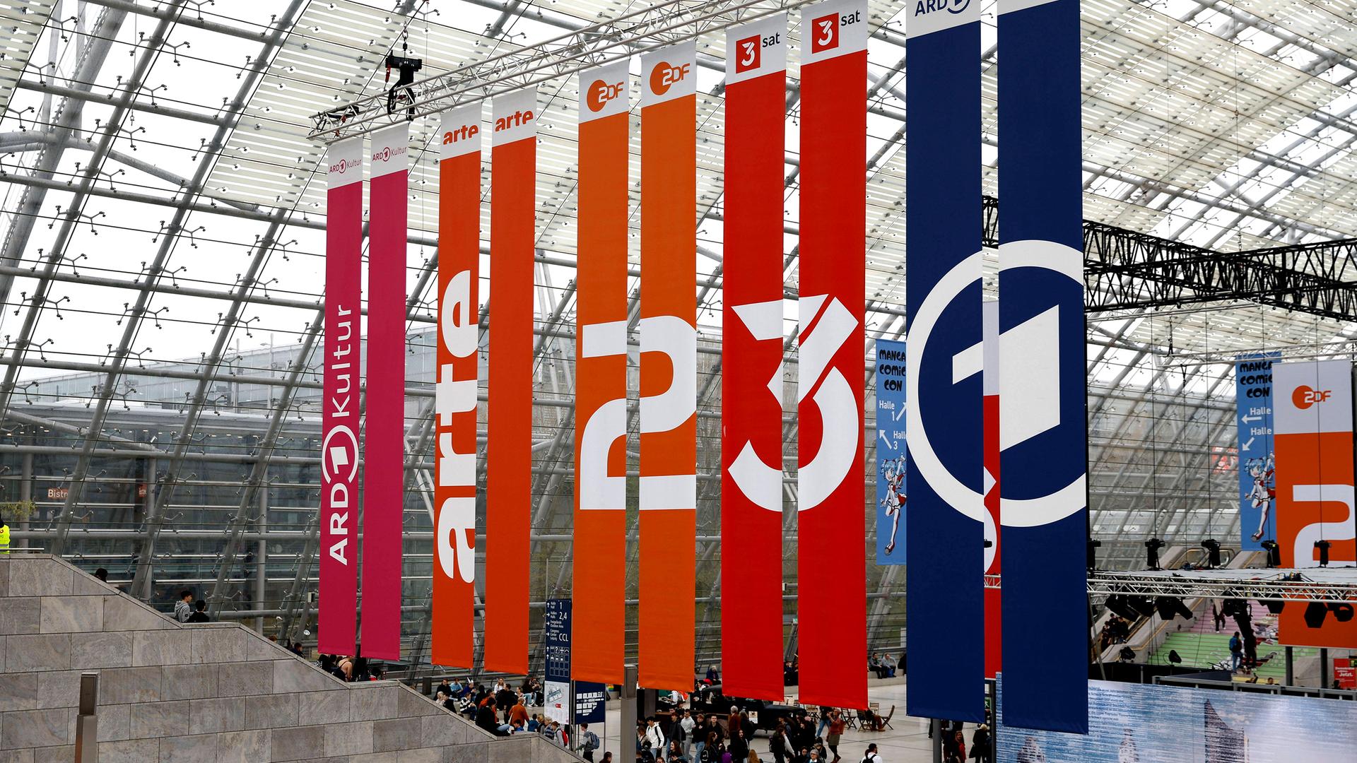 Vom Dach einer Glashalle hängen Banner mit den Logos von ARD, ZDF, 3sat und arte