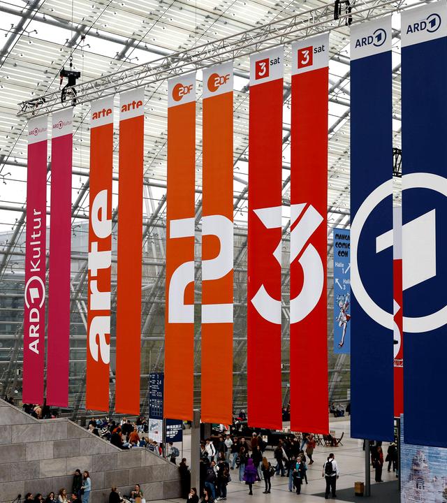 Vom Dach einer Glashalle hängen Banner mit den Logos von ARD, ZDF, 3sat und arte