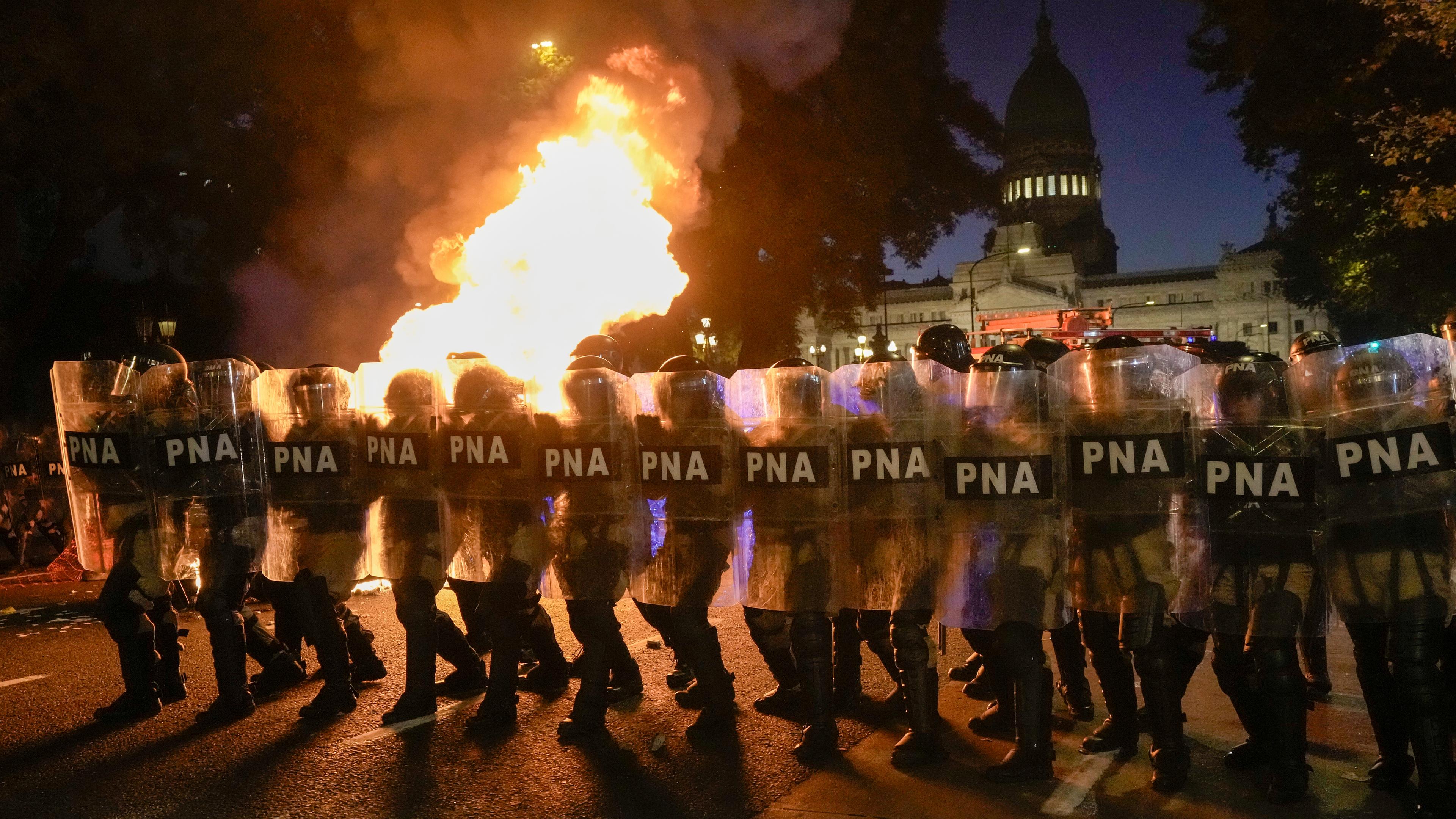Polizisten stehen Demonstranten in Buenos Aires gegenüber; im Hintergrund steigt Rauch auf.
