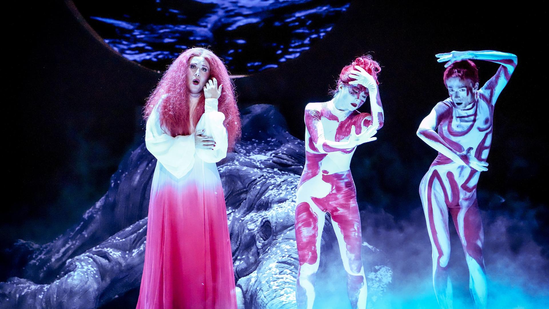 Das Bild zeigt eine Szene aus der Oper "Ariadne auf Naxos" von Richard Strauss, aufgeführt bei den Salzburger Festspielen 2026. Im Vordergrund sind drei Figuren zu sehen:   1. Eine Hauptdarstellerin, die längeres, lockiges, rötliches Haar trägt und ein langes, weißes Kleid mit einem sanften Farbverlauf von Weiß zu Rot anhat. Sie wirkt emotional und zieht ihre Hände an ihr Gesicht.  2. Zwei weitere Schauspielerinnen, die in hautengen Kostümen mit abstrahierten, roten und weißen Mustern gekleidet sind. Ihre Körper sind auffällig bemalt und wirken expressive, mit dynamischen Körperhaltungen.   Im Hintergrund ist eine stilisierte Kulisse mit einer gemäldeartigen Darstellung von Wasser oder einem Himmel zu sehen. Nebel umhüllt die Szene, was eine dramatische Atmosphäre schafft. Es ist eine eindrucksvolle, theatralische Inszenierung, die besondere Emotionen und Themen der Oper vermittelt.