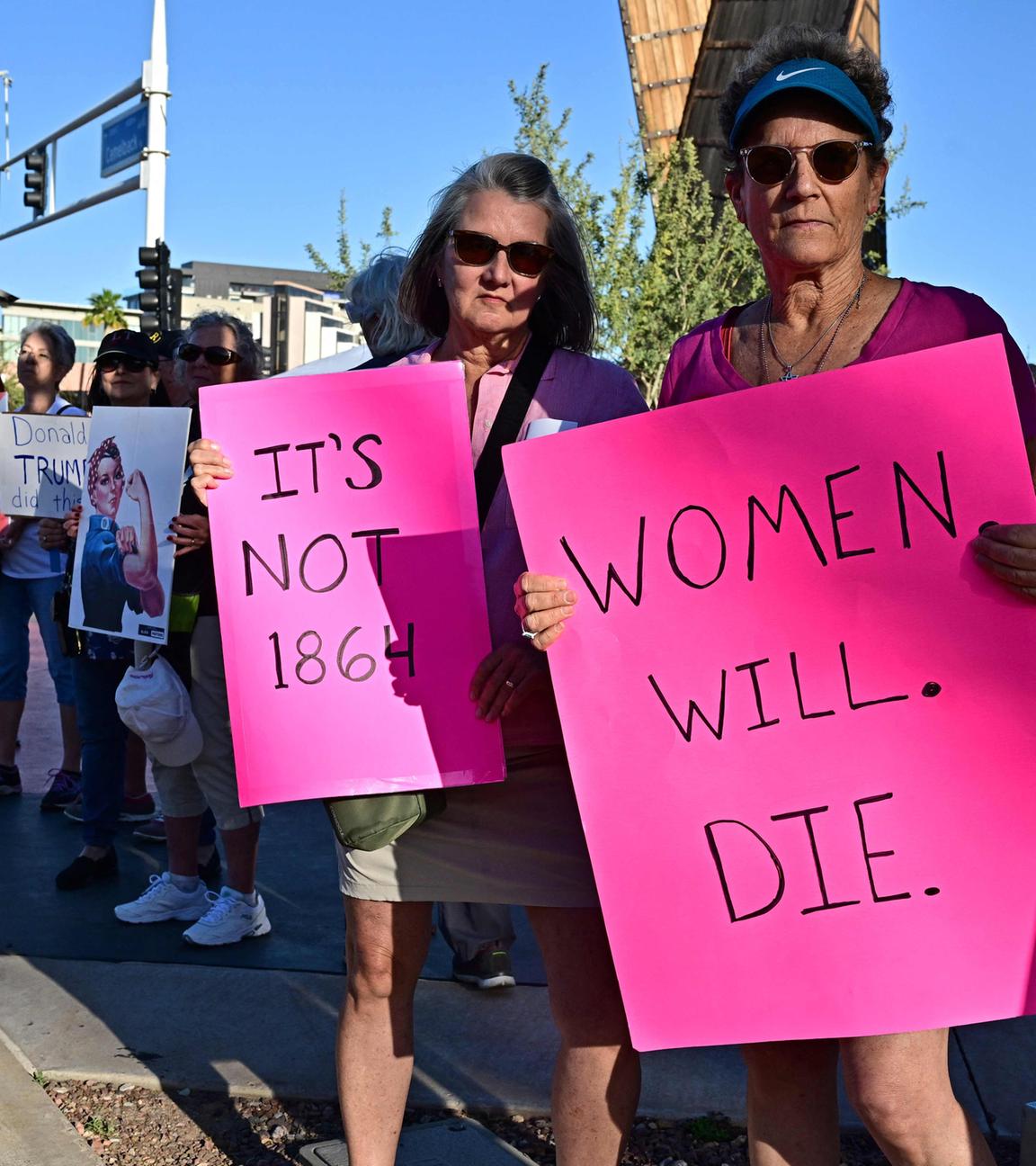 Am Straßenrand stehen Demonstranten mit Schildern, vorne sind zwei Frauen zu sehen, die pinke Schilder in die Kamera halten mit den Aufschriften "Frauen werden streben" und "Es ist nicht 1864".