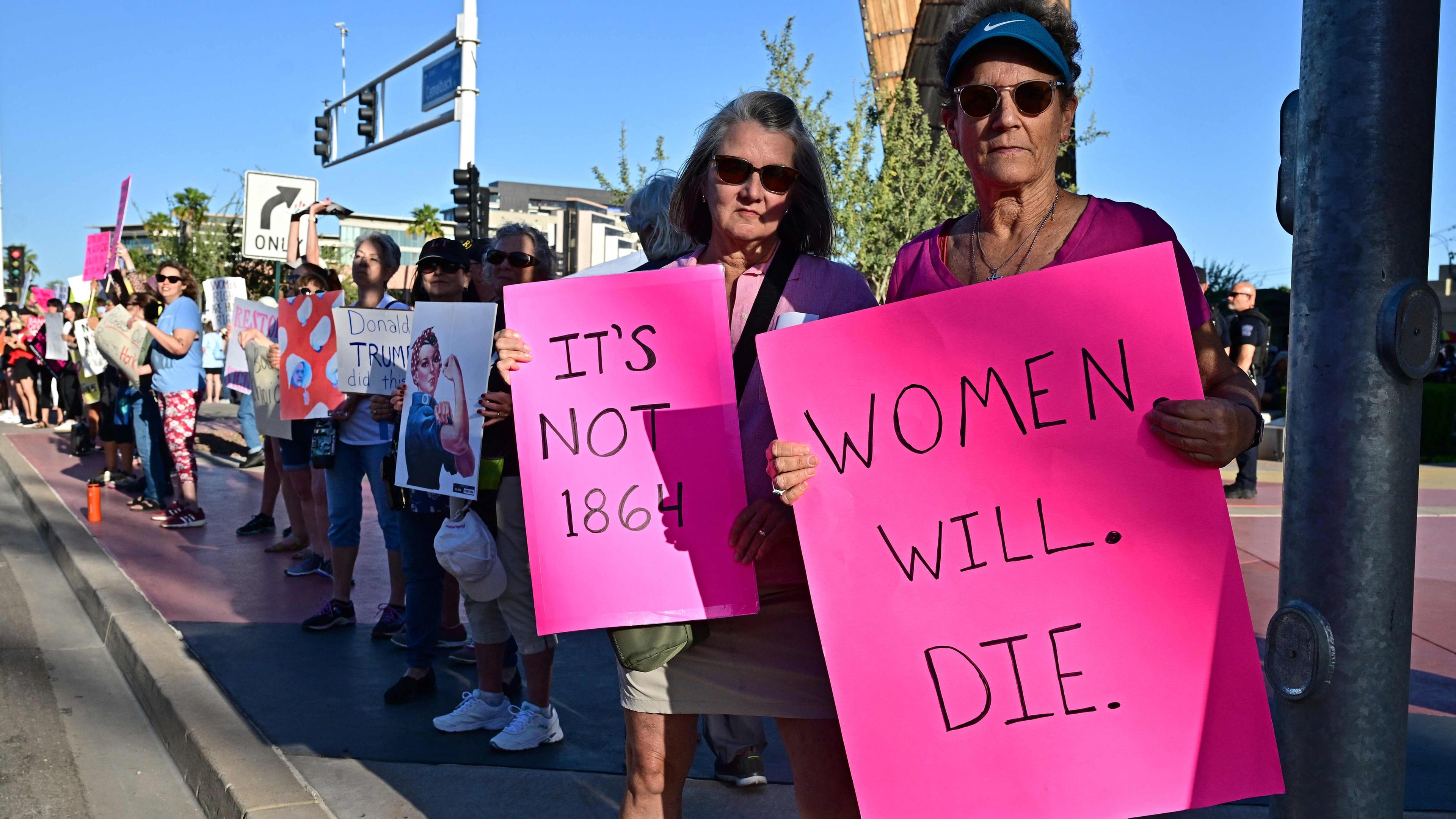 Am Straßenrand stehen Demonstranten mit Schildern, vorne sind zwei Frauen zu sehen, die pinke Schilder in die Kamera halten mit den Aufschriften "Frauen werden streben" und "Es ist nicht 1864".