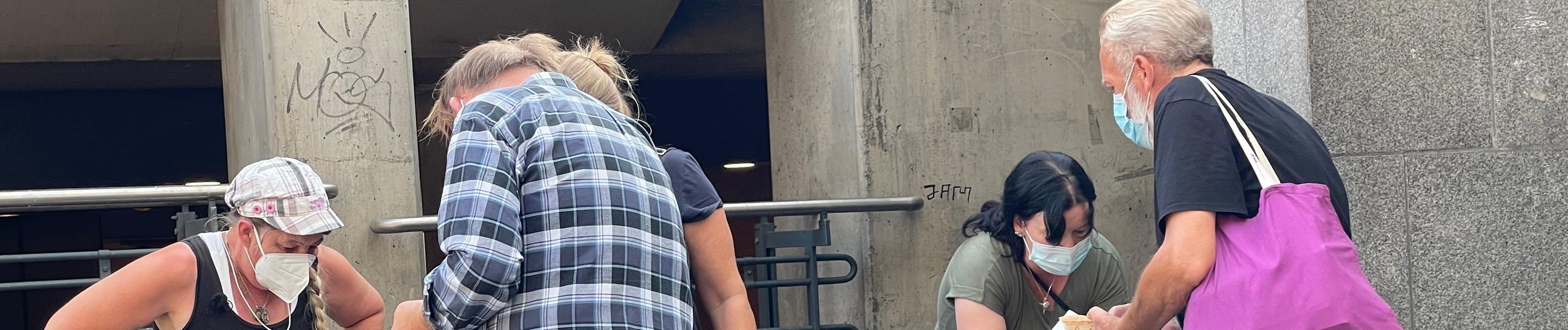 "Arm gegen Arm - Verteilungskampf am unteren Rand": Vor einem Betonbau wählen zwei Menschen Hilfsgüter aus zwei mobilen Behältern aus. Weitere kümmern sich um die Vergabe, alle tragen Corona-Schutzmasken.