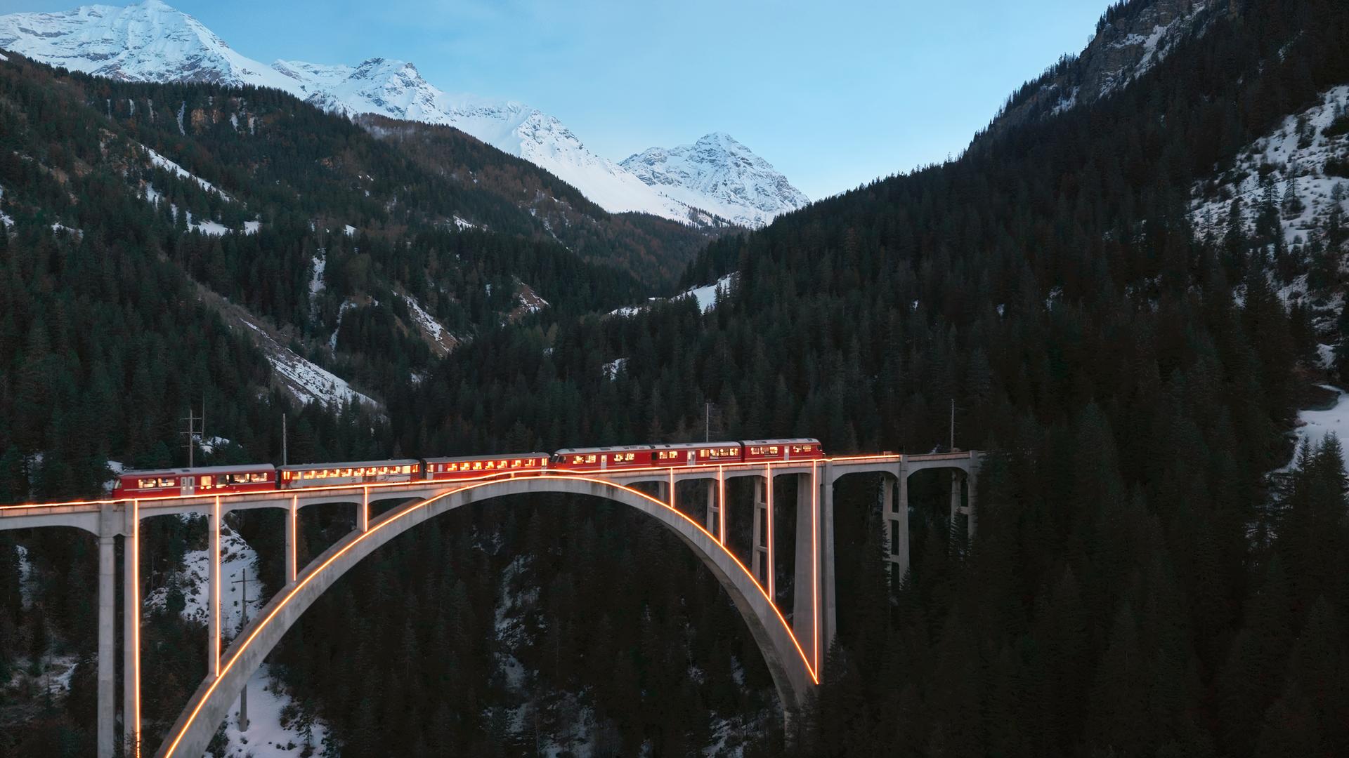 Ein roter Zug fährt in der Dämmerung über eine beleuchtete Brücke, umgeben von einer winterlichen Berglandschaft mit schneebedeckten Gipfeln.