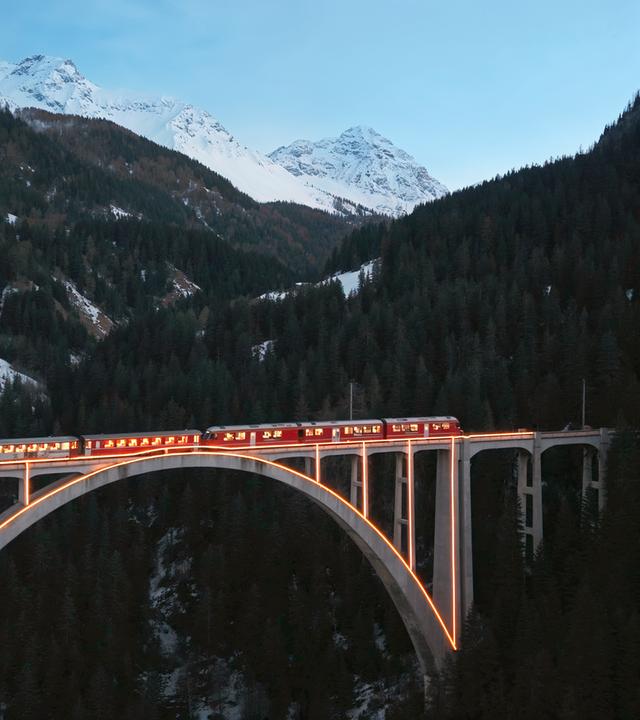 Ein roter Zug fährt in der Dämmerung über eine beleuchtete Brücke, umgeben von einer winterlichen Berglandschaft mit schneebedeckten Gipfeln.