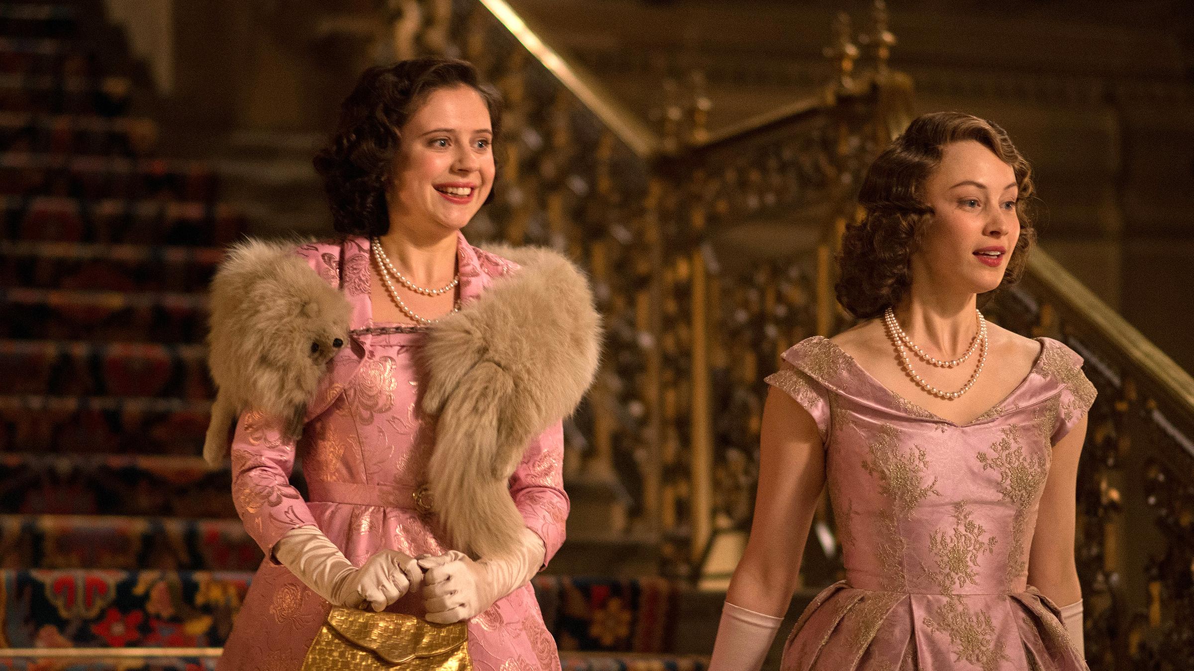 Die beiden Prinzessinnen Margaret (Bel Powley) und Elizabeth (Sarah Gadon) kommen nebeneinander eine prunkvolle Treppe herunter. Sie schauen sich lächelnd um. Beide tragen fast identische, elegante Kleider.